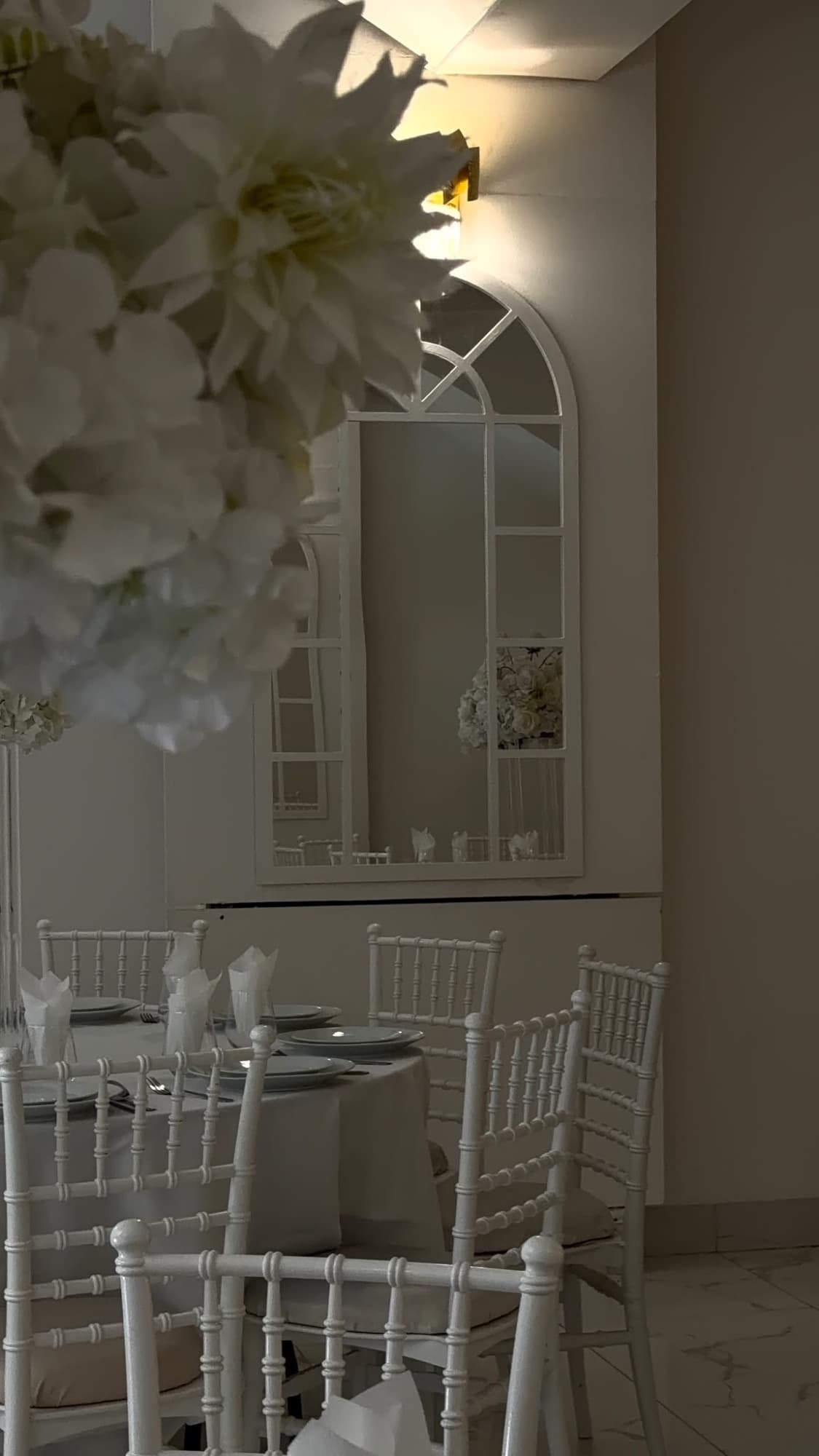 White chiavari chairs around a table, with flowers in front of an arched mirror